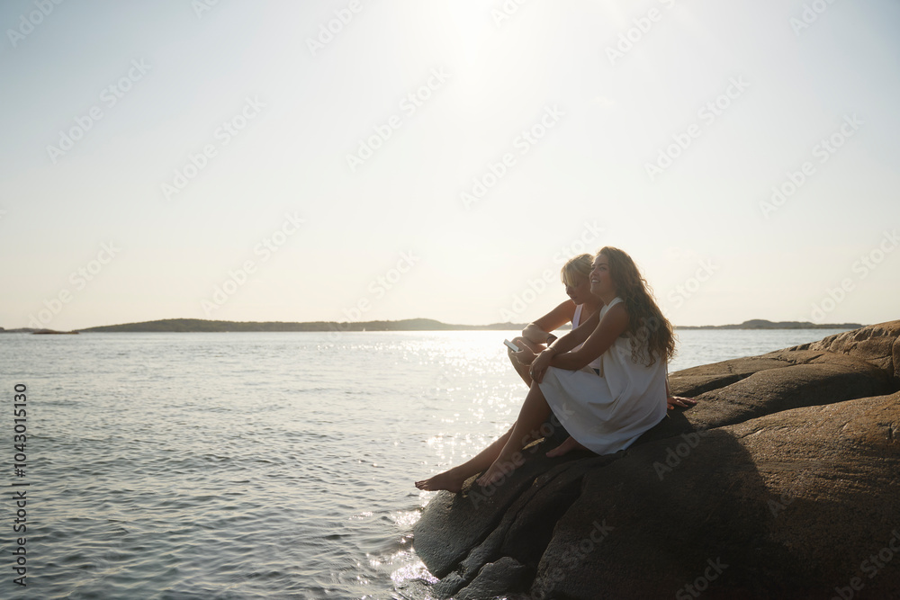 Full length of female friends sitting on rock against clear sky at lakeshore Full length of female friends sitting on rock against clear sky at lakeshore
