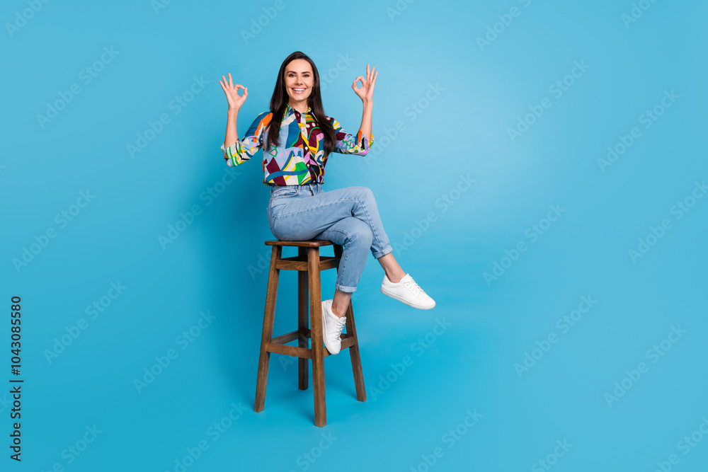 Full size photo of positive satisfied girl dressed print blouse jeans sit on chair showing okey approve isolated on blue color background Full size photo of positive satisfied girl dressed print blouse jeans sit on chair showing okey approve isolated on blue color background