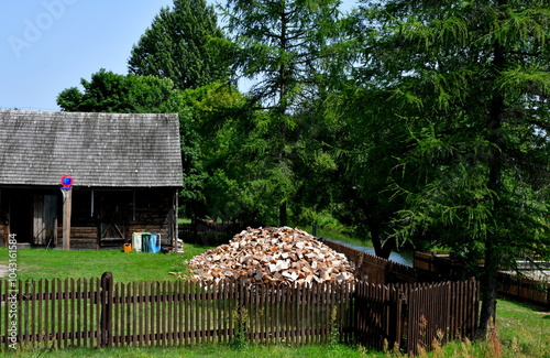 A view of an old wooden hut or shack surrounded with a wooden fence made out of planks and with a large pile of chopped wood nearby situated next to a lush forest or moor seen on a sunny summer day