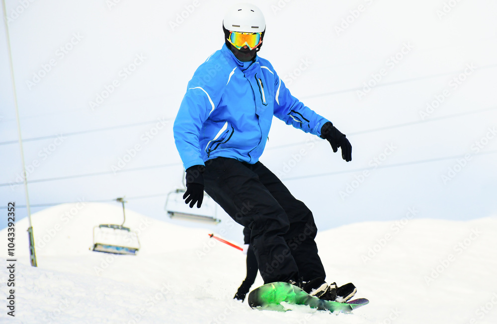 Naklejka premium close up casual snowboard rider in blue black outfit looks down to camera isolated in white snowy background. Winter vacation with sports mountains