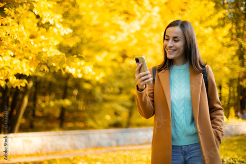 Young woman in a beige coat and light blue sweater is smiling while looking at her smartphone. She is walking in a sunlit autumn park with bright yellow foliage, enjoying a pleasant moment outdoors