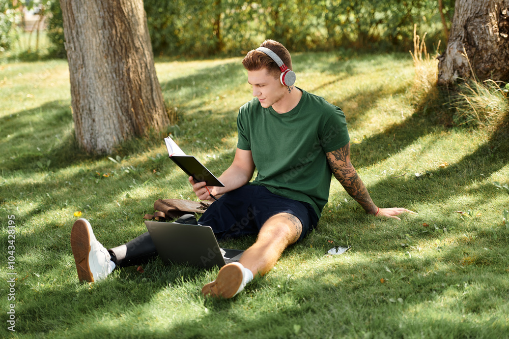 Fototapeta premium A young man relaxes on the grass, reading intently while wearing headphones and enjoying nature beauty.