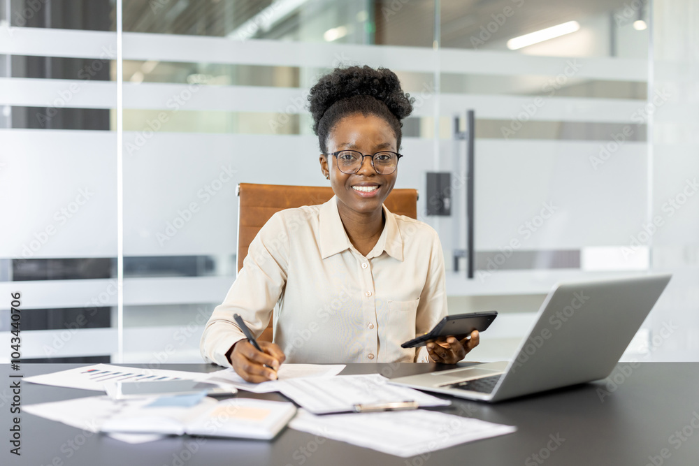 Fototapeta premium Confident African American businesswoman with calculator, phone, laptop, financial documents in office. Shows professionalism, finance expertise, productivity. Ideal for business, finance themes.