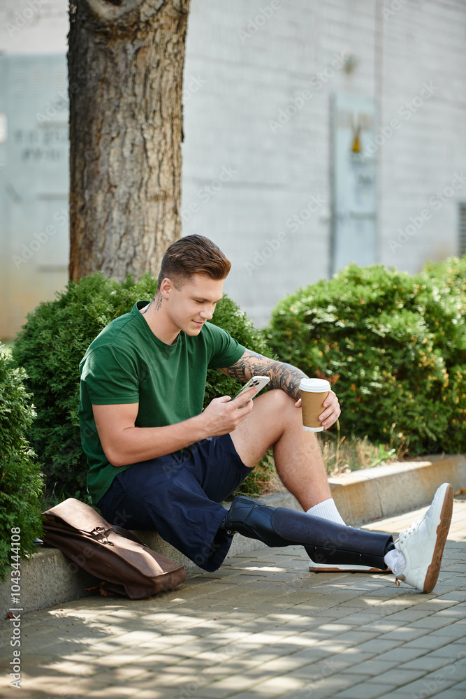 Fototapeta premium A young man with a prosthetic leg sits on the ground, sipping coffee and checking his phone in a sunny location.