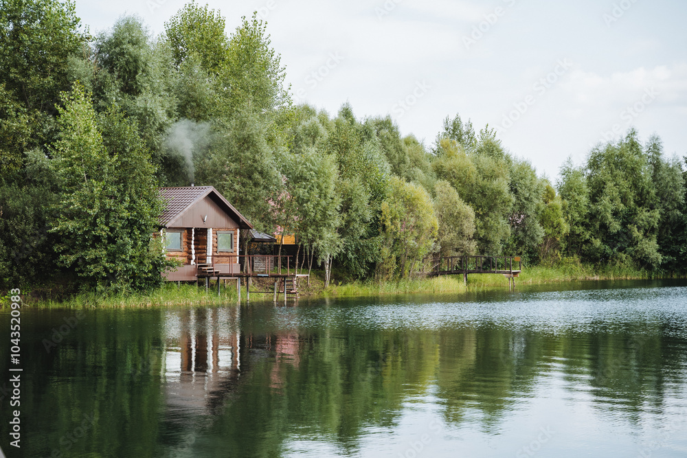 Obraz premium Nestled beside a serene lake, a quaint small wooden house stands gracefully, framed by lush green trees, creating a stunning view of the breathtaking natural landscape all around