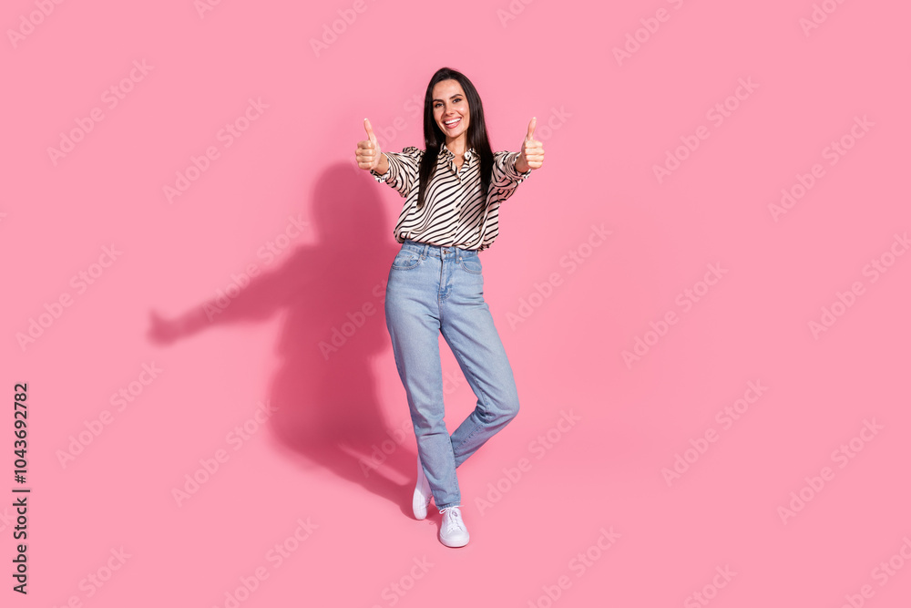 Confident young woman giving thumbs up standing against pink background in casual striped shirt and jeans mature female Confident young woman giving thumbs up standing against pink background in casual striped shirt and jeans mature female