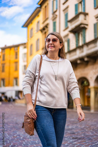 Portrait of beautiful middle-aged long-haired woman wearing jeans, beige sweater, sunglasses and leather handbag walking along street in european city on sunny autumn day. Front view