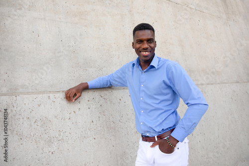 Portrait Of Handsome Smiling With Teeth African American Man wearing White Jeans and Blue Shirt, Brown Belt Outdoor, Concrete Wall on Background. Copy Space Fashionable Male, Ivorian. Horizontal photo