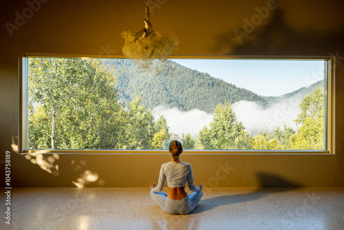 Fényképezés A woman sits in a meditative pose facing a large window, which frames a stunning view of mist-covered mountains and lush green trees