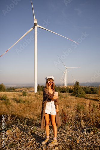 Stylish woman takes a selfie in front of massive wind turbines, embracing the intersection of modern fashion and sustainable energy in a scenic rural landscape