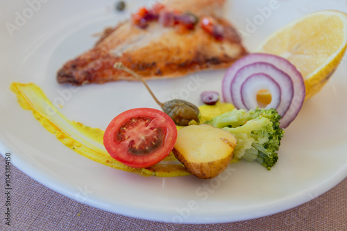 Obraz na płótnie Grilled sea bass in a white porcelain plate. Garnish: lemon, onion, tomato, capers, broccoli, potatoes, carrots. w salonie