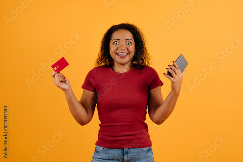 Emotional cheerful attractive young black woman with bushy hair and teeth braces holding red bank card and cell phone in her hands, purchasing online, yellow studio background. High quality photo photo