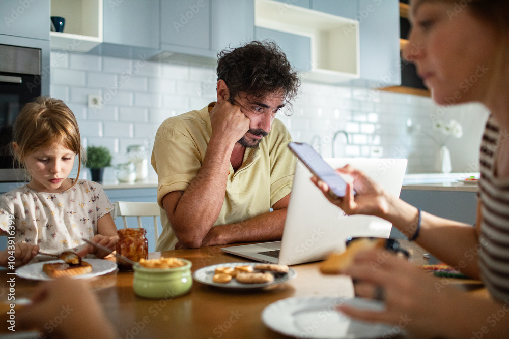 Father working on laptop while family has breakfast in kitchen Father working on laptop while family has breakfast in kitchen
