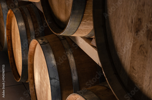 A close-up of several wooden wine barrels stacked in a cellar. The barrels are made of dark brown wood with silver metal hoops. The image has a warm, rustic feel.