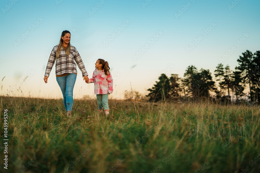 A mother and daughter walking hand in hand through a grassy field at sunset, with trees in the background. A mother and daughter walking hand in hand through a grassy field at sunset, with trees in the background.