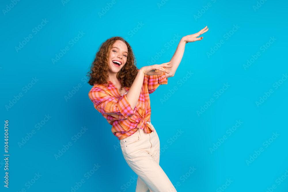 Naklejka premium Cheerful young woman with curly red hair gesturing and smiling in a bright outfit against a blue background.