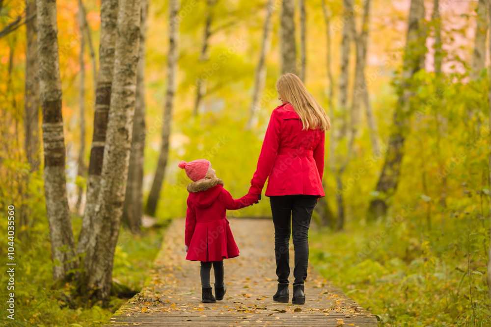 Happy little daughter and young adult mother in red coats walking on trail at birch tree forest. Spending time together in beautiful colorful autumn day. Enjoying stroll. Back view. Happy little daughter and young adult mother in red coats walking on trail at birch tree forest. Spending time together in beautiful colorful autumn day. Enjoying stroll. Back view.