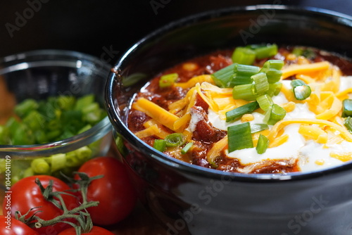 Bowl of fresh hot steaming chili in a black bowl with sour cream, cheese and chives.  Classic bowl of spicy chili for dinner.
