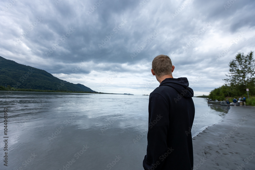 Fototapeta premium An explorer stands quietly on the bank of Alaska’s Knik River, gazing at the serene, icy waters under the soft glow of a cold, crisp morning.