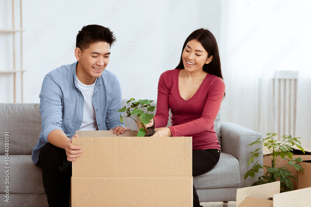 Relocation. Joyful Asian Couple Moving House And Packing Plant In Cardboard Box Sitting On Sofa At Home. Copyspace Relocation. Joyful Asian Couple Moving House And Packing Plant In Cardboard Box Sitting On Sofa At Home. Copyspace
