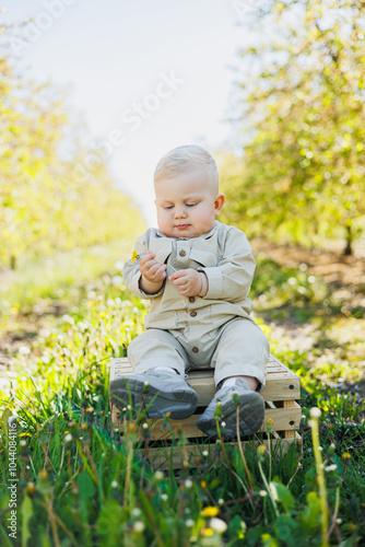 A cute baby boy is sitting on the grass in the park and smiling. A stylish baby in a beige romper.