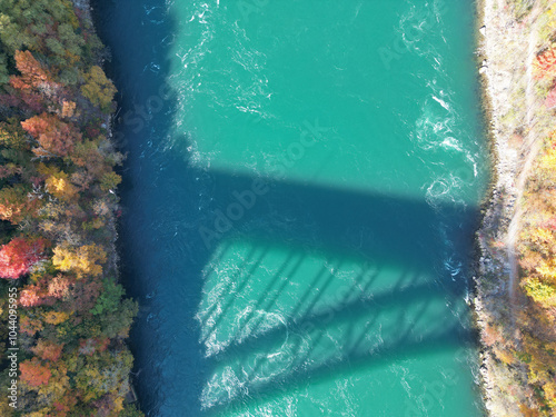 Aerial of the shadow of the international border crossing Queenston-Lewiston bridge over the Niagara River. Shot during an autumn afternoon with colorful fall foliage trees in October, 2024.