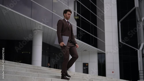 Confident businessman in suit descending office building stairs with laptop in hand, showcasing professional demeanor. Handsome sbusiness man successful walking down stairs of modern office building. photo