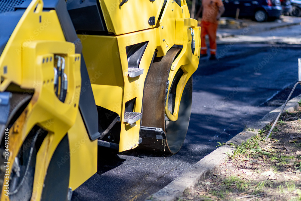 Construction workers using a yellow asphalt roller during road paving at a city site on a sunny day in early autumn Construction workers using a yellow asphalt roller during road paving at a city site on a sunny day in early autumn