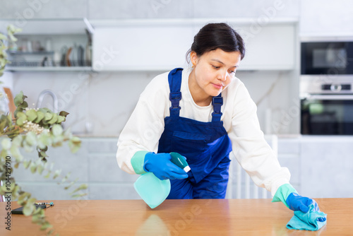 Portrait of interested diligent young female janitor making modern office kitchen shine, dusting furniture with cleaning spay..