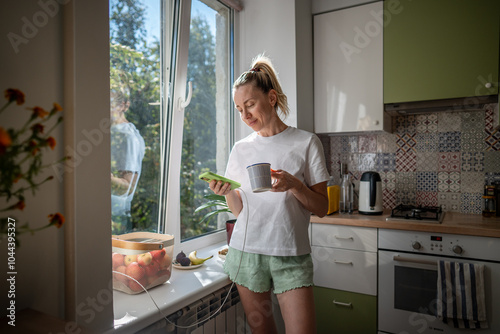 Картината върху платно Calm smiling woman with coffee mug and phone unwinds in kitchen