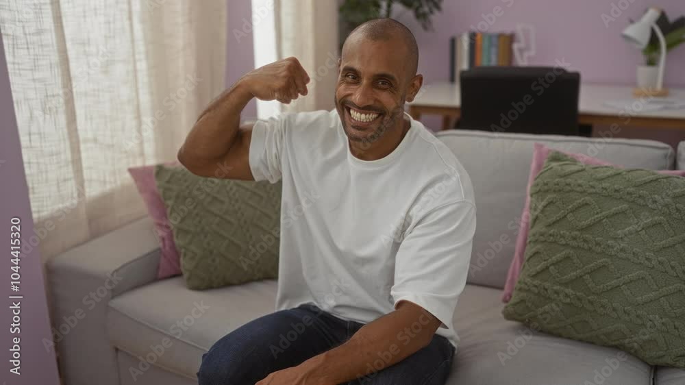Young man flexing muscles and smiling while sitting on a sofa in a cozy living room setting with a desk behind him showing a relaxed home environment Young man flexing muscles and smiling while sitting on a sofa in a cozy living room setting with a desk behind him showing a relaxed home environment