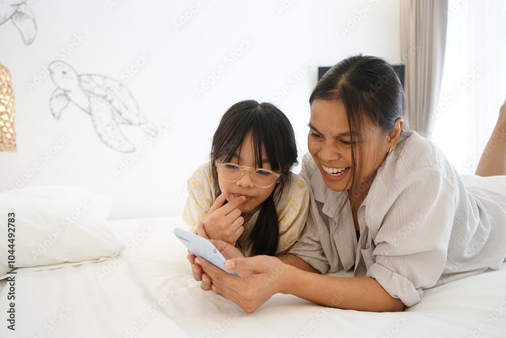 Close up caring Asian mother and little girl using smartphone together, lying on cozy bed in bedroom, family enjoying leisure time with gadget at home Close up caring Asian mother and little girl using smartphone together, lying on cozy bed in bedroom, family enjoying leisure time with gadget at home