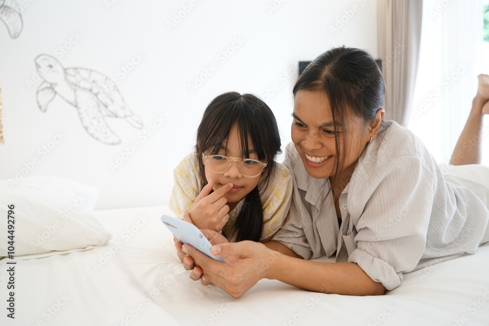 Close up caring Asian mother and little girl using smartphone together, lying on cozy bed in bedroom, family enjoying leisure time with gadget at home Close up caring Asian mother and little girl using smartphone together, lying on cozy bed in bedroom, family enjoying leisure time with gadget at home