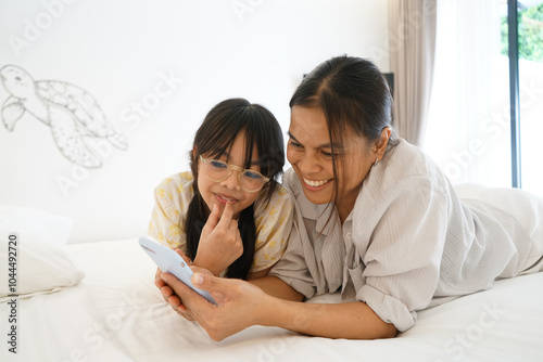 Close up caring Asian mother and little girl using smartphone together, lying on cozy bed in bedroom, family enjoying leisure time with gadget at home