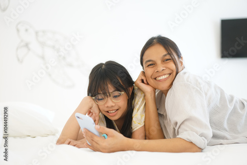 Close up caring Asian mother and little girl using smartphone together, lying on cozy bed in bedroom, family enjoying leisure time with gadget at home