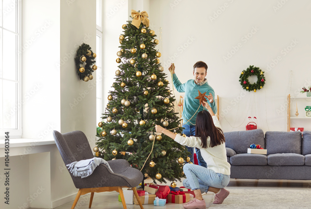 Happy joyful smiling young couple in love decorating Christmas fir tree together standing in festive living room at home preparing for New Year celebration. Winter holidays concept. Happy joyful smiling young couple in love decorating Christmas fir tree together standing in festive living room at home preparing for New Year celebration. Winter holidays concept.