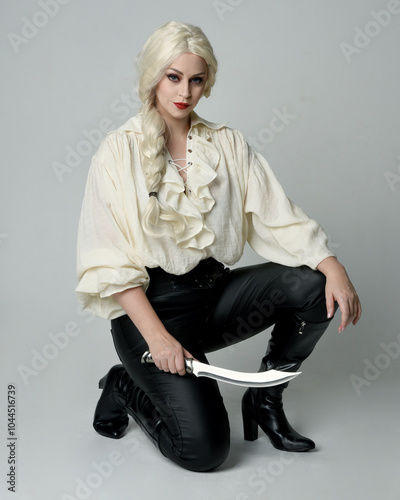 full length portrait of beautiful female model, wearing vintage gothic fantasy ruffle blouse white shirt, leather pants, blonde hair. holding knife weapon. sitting pose, isolated on studio background.