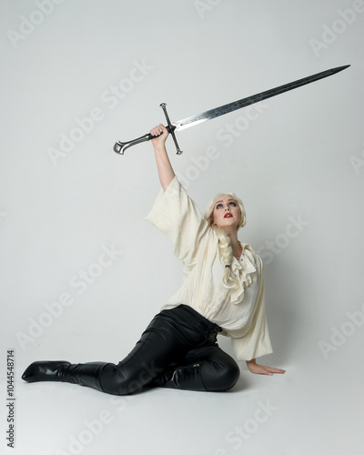 Bild auf Leinwand Full length portrait of female model wearing vintage gothic fantasy blouse, leather pants and white blonde hair, kneeling holding sword weapon, isolated figure on studio background
