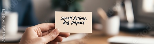 Close-up of a hand holding a motivational card with the message 'Small Actions, Big Impact' in an office environment, inspiring productivity.