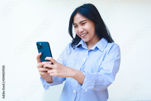 excited asian woman using smartphone apps wearing blue striped shirt isolated on white background, playful female surfing in social media