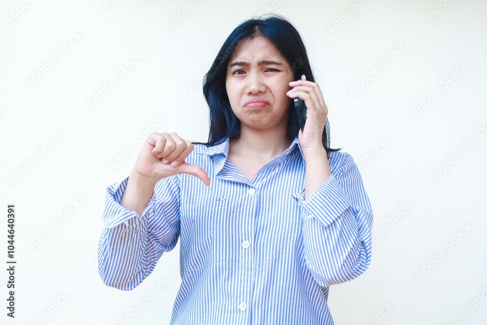 sad asian woman talking on smartphone showing thumbs down feeling upset wearing striped shirt isolated on whtie background sad asian woman talking on smartphone showing thumbs down feeling upset wearing striped shirt isolated on whtie background