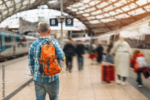 A traveler stands with an orange backpack at a bustling train station during a sunny afternoon
