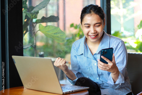 success asian woman looking at smartphone and raising fist celebrate victory and achievement while working on laptop in cafe