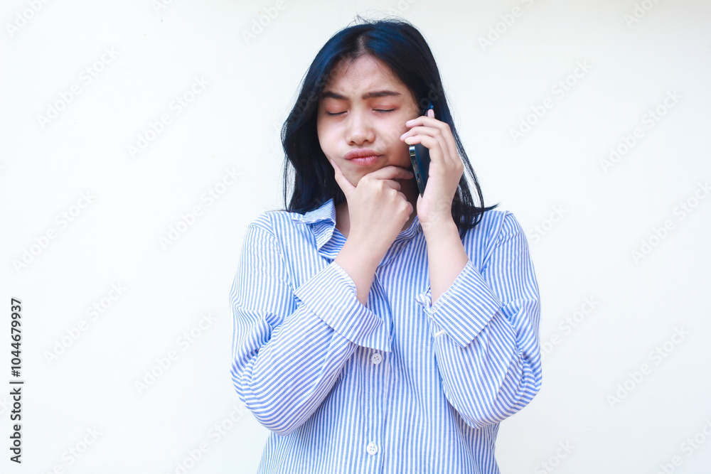 thoughtful asian woman using smartphone with hands on chin thinking problem wearing blue striped casual shirt standing over isolated white background, look away thoughtful asian woman using smartphone with hands on chin thinking problem wearing blue striped casual shirt standing over isolated white background, look away