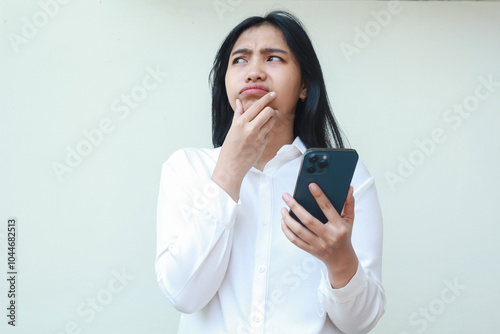 curious asian young business woman looking up wondering and thinking while using mobile phone app with hand on chind, surfing internet, wear white formal suit standing over white background