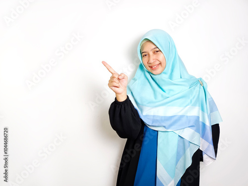 East asian indonesian muslim woman with hijab gamis smiling pointing finger to copy space isolated on white background.
 photo