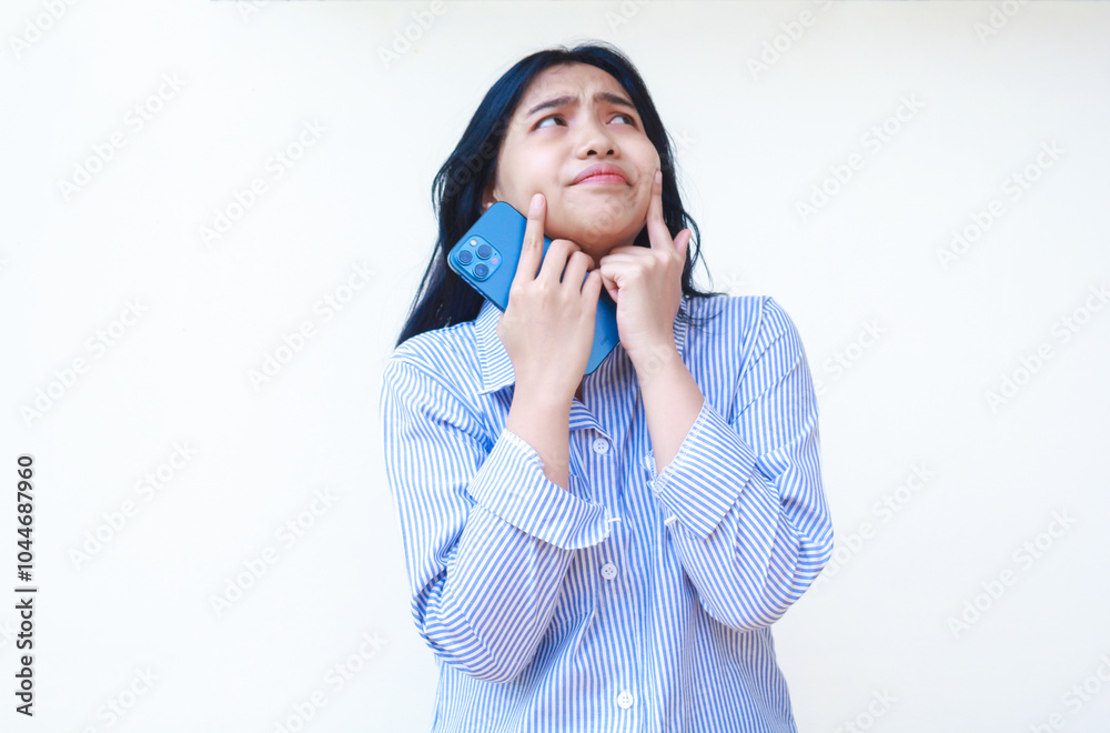 thoughtful asian woman using smartphone with hands on cheek thinking problem wearing blue striped casual shirt standing over isolated white background, look away thoughtful asian woman using smartphone with hands on cheek thinking problem wearing blue striped casual shirt standing over isolated white background, look away