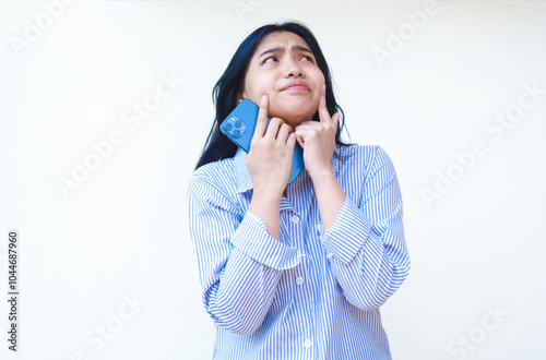 thoughtful asian woman using smartphone with hands on cheek thinking problem wearing blue striped casual shirt standing over isolated white background, look away