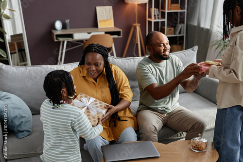 Family sharing joyful moment during special occasion in living room setting. Child presenting gift to mother while father observing with smile. Cozy, warm atmosphere capturing happiness photo