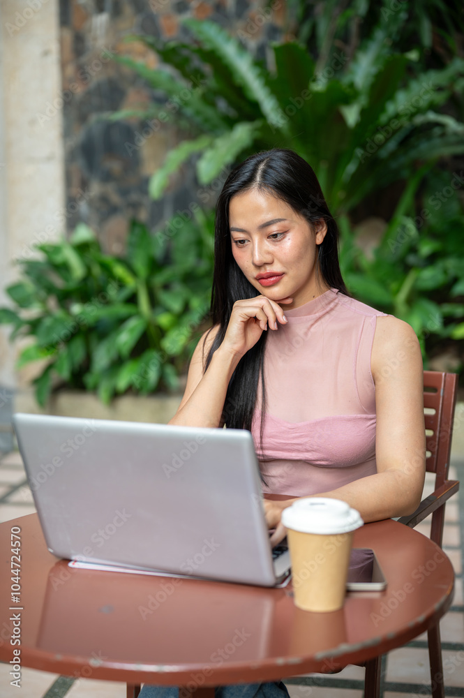 A beautiful, focused Asian woman is working remotely, working on her laptop at an outdoor cafe. A beautiful, focused Asian woman is working remotely, working on her laptop at an outdoor cafe.